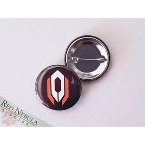 May include: A black and orange button with a white logo. The logo is a stylized letter O with two vertical lines on either side. The button is 1 inch in diameter.