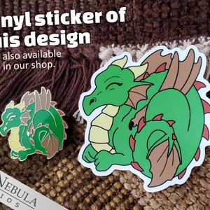 May include: A green dragon sticker and pin with brown and red accents. The sticker and pin feature the text "Vinyl sticker of this design also available in our shop."