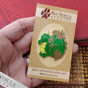 May include: A green and brown dragon enamel pin on a tan card. The dragon is curled up with a yellow belly and red accents. The card has the text "Red Nebula Studios" and website information.