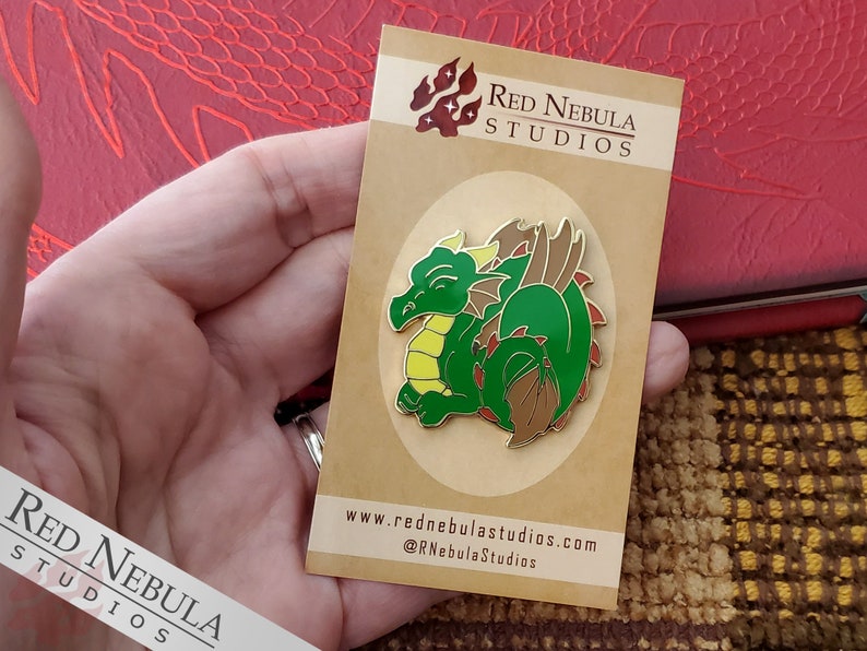 May include: A green and brown dragon enamel pin on a tan card. The dragon is curled up with a yellow belly and red accents. The card has the text "Red Nebula Studios" and website information.