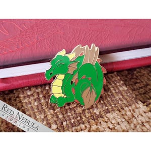 May include: A green dragon enamel pin with yellow and brown accents. The dragon is in a relaxed pose with its eyes closed. The pin is set against a red book and a textured brown surface. The text "Red Nebula Studios" is visible.