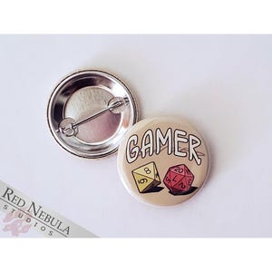 May include: A round button with a beige background featuring the word "GAMER" in white text. Two dice are shown below the text, one red and one yellow.