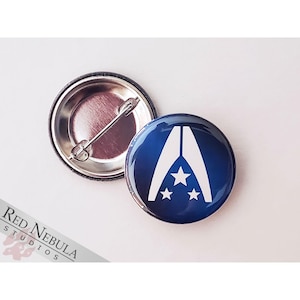 May include: A blue and white button with a white stylized letter A and three white stars. The button is a pin-back style.