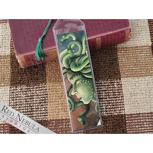 May include: A bookmark featuring a detailed illustration of a woman with snakes for hair. The woman's face is partially obscured by the snakes, and her eyes are closed. The snakes are green and brown, and the background is a reddish-brown color.