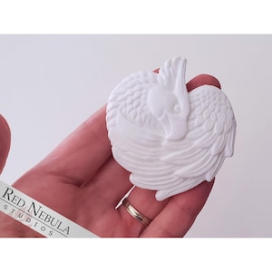 May include: White ceramic sculpture of a bird with its wings folded. The bird's head is tucked into its wings. The sculpture is detailed with feathers.
