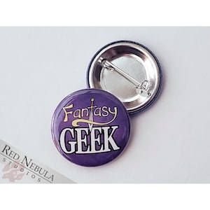 May include: A purple button with the words "Fantasy GEEK" in white and gold lettering. The button has a silver pin back and a dark blue rim. The button is a round shape.