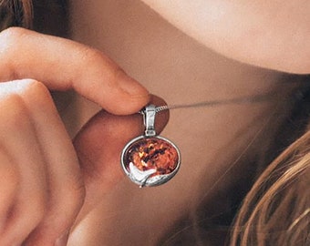 May include: A silver necklace with a pendant featuring a round, orange amber stone. The pendant is held by a woman's hand.
