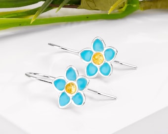 May include: A pair of silver-tone earrings featuring a flower design. Each earring has a light blue enamel flower with a yellow center. The earrings are displayed on a white surface with green foliage in the background.