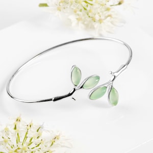 May include: A silver bangle bracelet with a delicate branch design featuring light green gemstone leaves.
