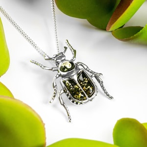 May include: A silver necklace with a beetle pendant featuring a green amber stone. The pendant is detailed with intricate silver work and the amber stone is a deep green colour.
