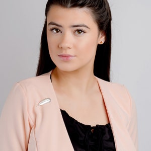 May include: A woman with long dark hair wearing a blush pink blazer with a silver feather brooch.