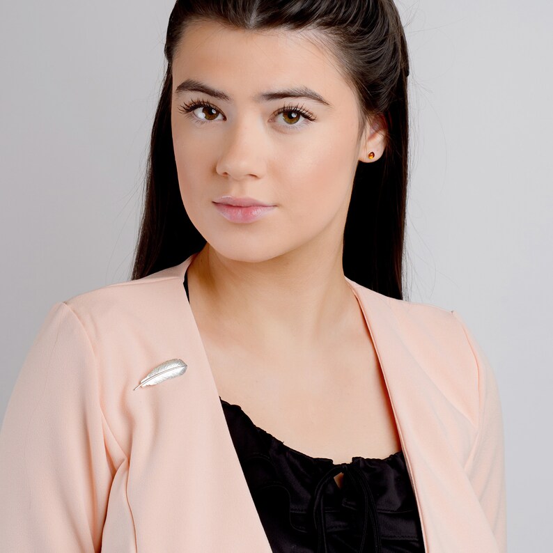 May include: A woman with long dark hair wearing a blush pink blazer with a silver feather brooch.
