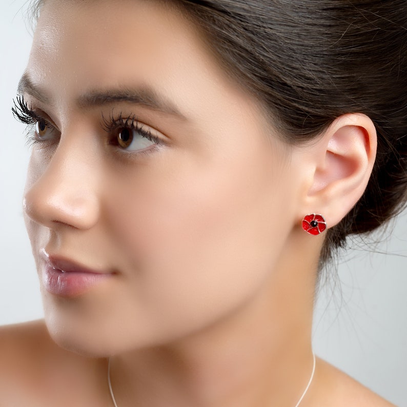 K&ouml;nnte beinhalten: Ein Paar silberne Ohrstecker mit einem roten Mohnblumen-Design. Die Ohrringe sind klein und zart, und die Mohnblumen sind mit schwarzen Zentren detailliert.
