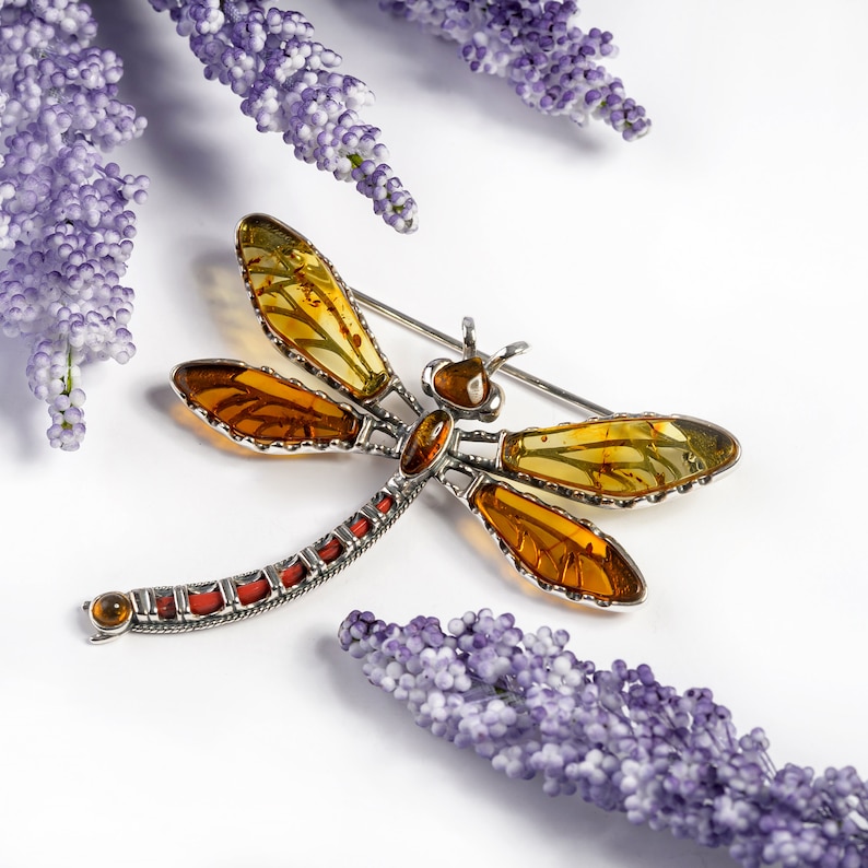May include: A silver dragonfly brooch with amber wings. The wings are a mix of yellow and orange amber. The brooch is set against a white background with lavender flowers.