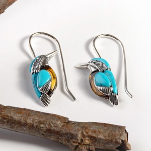 May include: A pair of silver dangle earrings featuring a turquoise and amber bird design. The earrings have a hook closure.
