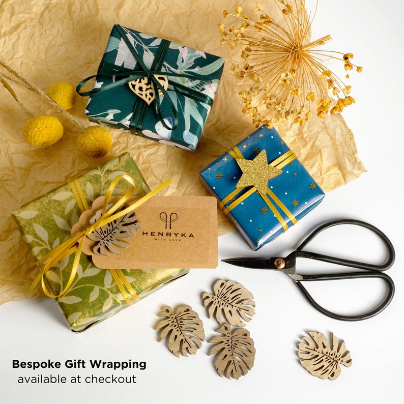 K&ouml;nnte beinhalten: Drei Geschenkboxen, die in verschiedenen gemusterten Papieren verpackt sind. Die Boxen befinden sich auf einem braunen Papieruntergrund. Die Boxen sind mit B&auml;ndern, einem Stern und einem Blatt verziert. Der Text "HENRYKA with love" befindet sich auf einem braunen Etikett, das an einer der Boxen befestigt ist. Der Text "Individuelle Geschenkverpackung an der Kasse erh&auml;ltlich" befindet sich am unteren Rand des Bildes.