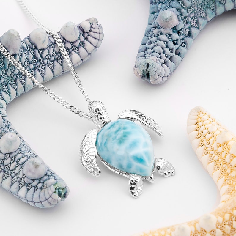 Op de afbeelding: Een zilveren schildpad hanger ketting met een blauwe en witte iriserende edelsteen. De ketting wordt getoond op een witte achtergrond met twee zeesterren.
