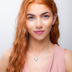 Op de afbeelding: Een vrouw met lang, golvend, rood haar draagt een lichtroze top zonder mouwen. Ze draagt een zilveren ketting met een turquoise zeeschildpad hanger.