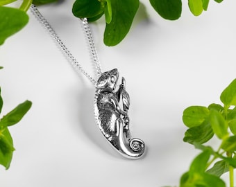 Könnte beinhalten: Eine silberne Chamäleon-Anhänger-Halskette an einer silbernen Kette. Das Chamäleon ist in einer Kletterpose dargestellt, wobei sein Schwanz um einen Ast gewickelt ist.