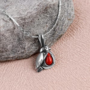 May include: A silver bird pendant with a red, teardrop-shaped stone. The pendant hangs from a delicate silver chain. The bird has detailed wings and a small beak. The pendant is displayed on a textured grey rock.