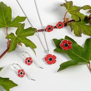 K&ouml;nnte beinhalten: Ein Set aus silbernem Mohnblumen-Schmuck mit einer Halskette, Ohrringen und einem Ring. Die Mohnblumen sind rot mit schwarzen Zentren und liegen auf einem wei&szlig;en Hintergrund mit gr&uuml;nen Bl&auml;ttern.