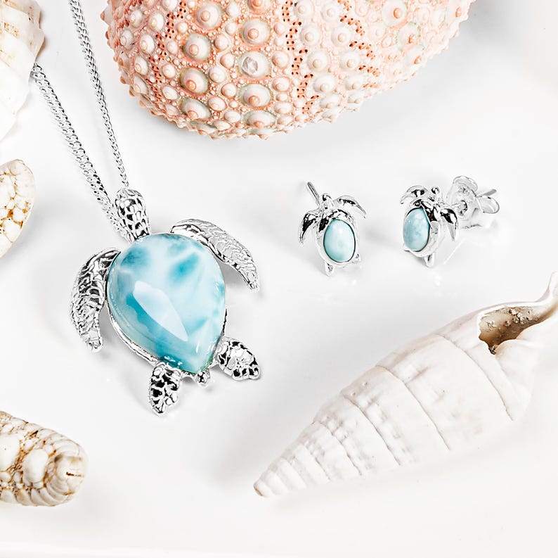 Op de afbeelding: Een zilveren schildpad ketting en oorbellen set met een blauwe en witte edelsteen hanger. De sieraden worden getoond op een wit oppervlak met schelpen.