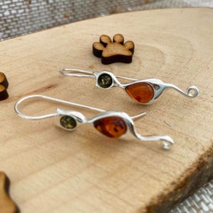Op de afbeelding: Een paar zilveren oorhangers met amber en groene stenen. De oorbellen hebben een uniek, gebogen ontwerp.