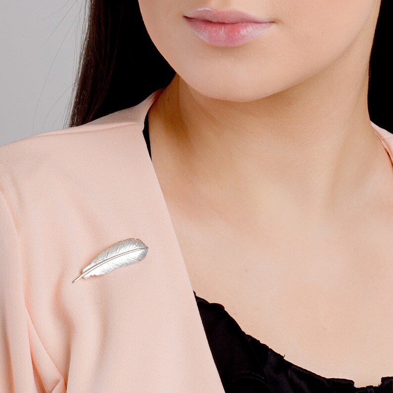 May include: A silver feather brooch pinned to a pink blazer. The brooch is a simple design with a detailed feather shape.