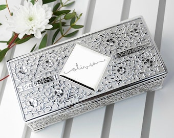 May include: A rectangular silver-toned metal box featuring an intricate embossed floral and vine pattern across the lid. A smooth, diamond-shaped central panel is engraved with the name olivia in a cursive script font. The box has ornate sides.