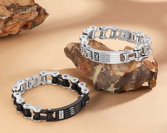 May include: Two silver chain bracelets with a black metal plate that says "DAD" and has a list of words on it. The bracelets are on a brown rock surface.