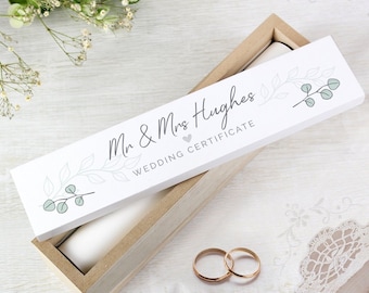 May include: White wooden box with a lid that says "Mr. & Mrs. Hughes Wedding Certificate" with a small heart and a leafy design. The box is open and two gold wedding bands are inside.