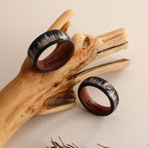 May include: Two black and silver wood rings with a forest design and the name "Emma" engraved on the inside of each ring.