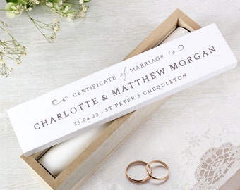 May include: A wooden box with a white lid, displaying the text "CERTIFICATE of MARRIAGE" and the names "CHARLOTTE & MATTHEW MORGAN". Two gold wedding rings are in the foreground. The date "25.04.23" is also visible.