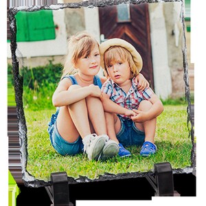 May include: Two children, a girl and a boy, are sitting on a grassy lawn in front of a house. The girl is wearing a blue and white dress and the boy is wearing a checked shirt and a straw hat. They are both smiling and looking at the camera.