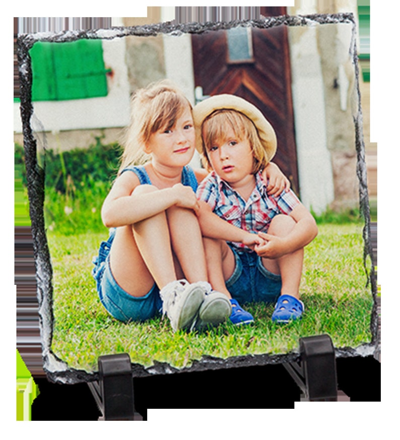 May include: Two children, a girl and a boy, are sitting on a grassy lawn in front of a house. The girl is wearing a blue and white dress and the boy is wearing a checked shirt and a straw hat. They are both smiling and looking at the camera.