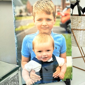 May include: A photo slate featuring a portrait of two young boys. The older boy is wearing a blue shirt and is holding the younger boy, who is wearing a blue and brown overall.