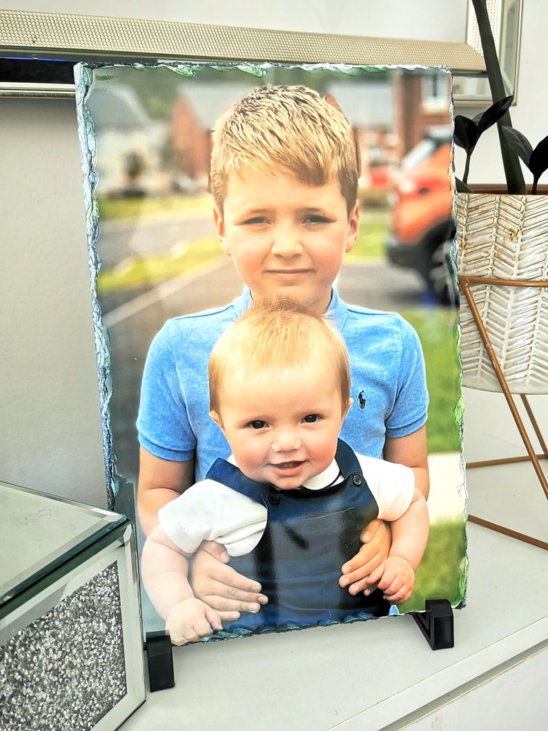 May include: A photo slate featuring a portrait of two young boys. The older boy is wearing a blue shirt and is holding the younger boy, who is wearing a blue and brown overall.