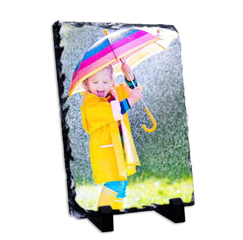 May include: A child wearing a yellow raincoat and blue trousers holds a colourful umbrella while standing in the rain. The child is smiling and sticking their tongue out.