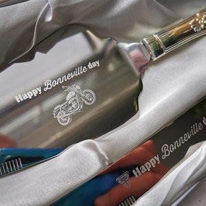 May include: A silver cake server and knife set with engraved text "Happy Bonneville day" and a motorcycle illustration. The server has a decorative handle.