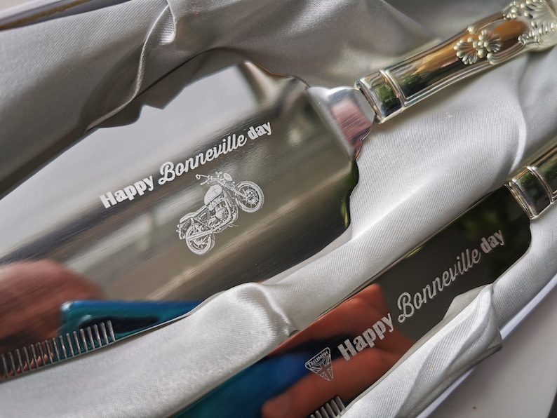 May include: A silver cake server and knife set with engraved text "Happy Bonneville day" and a motorcycle illustration. The server has a decorative handle.