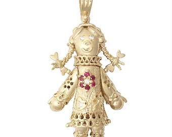May include: A gold-colored doll pendant with a loop for a chain. The doll has braided pigtails, a decorative collar, and a dress with a floral design. The dress features a flower with red and white gemstones. The doll's eyes are small gemstones.