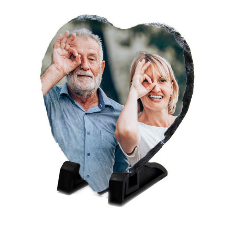 May include: A heart-shaped slate photo frame with a black stand, featuring a photo of an older couple smiling and making silly faces with their hands.