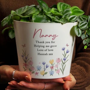 May include: A white plant pot with a floral design and the word "Nanny" in cursive. The pot has a message that reads "Thank you for Helping me grow Love of love Hannah xxx". The pot is filled with green leafy plants.