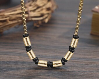 May include: A gold-colored necklace with a chain and cylindrical beads. Each bead is engraved with a name: Moletema, Brenda, Maddie, Paula, Melina, and Oakley. The beads are separated by black spacers. The necklace is displayed on a wooden surface.