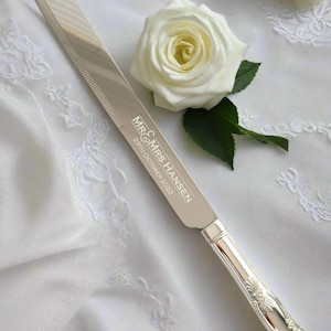 May include: Silver cake knife with an ornate handle and a serrated blade. The knife is engraved with "MR & MRS HANSEN 29th OCTOBER 2022".