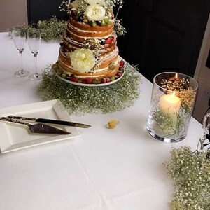 May include: A white wedding cake with multiple tiers decorated with fresh flowers, fruit, and baby's breath. The cake is on a white cake stand and is surrounded by baby's breath. There are two champagne flutes and a candle in a glass vase on a white tablecloth.