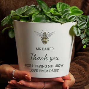 May include: A white plant pot featuring a bee design and the words "MR BAKER Thank you FOR HELPING ME GROW LOVE FROM DAISY". The pot is filled with green foliage.