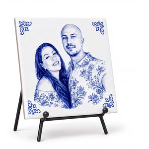 May include: A square white ceramic tile featuring a blue ink portrait of a couple. The tile has decorative blue corner flourishes and rests on a black metal display stand. The couple is depicted in a detailed sketch style with floral patterns.