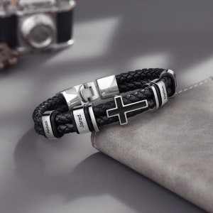 May include: Black leather bracelet with silver metal accents and a black cross charm. The bracelet has three engraved names: Abbas, Jared, and Daimon.