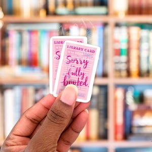 Może przedstawiać: Dwa r&oacute;żowe naklejki w kształcie karty bibliotecznej z tekstem "Przepraszam, jestem w pełni zarezerwowany". Naklejki są trzymane w dłoni na tle rozmytych regał&oacute;w z książkami.